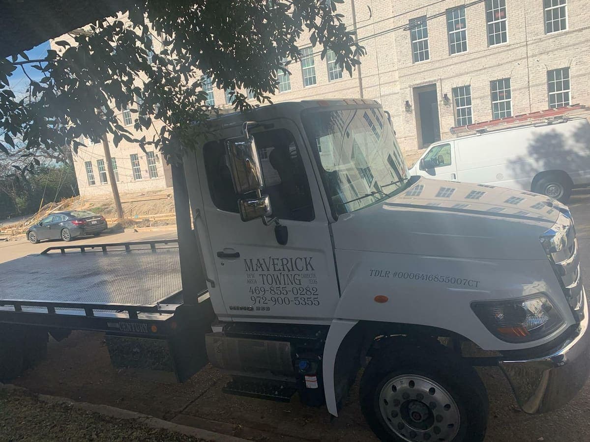 Maverick Towing white Hino flatbed tow truck parked under a tree beside a Dallas office building