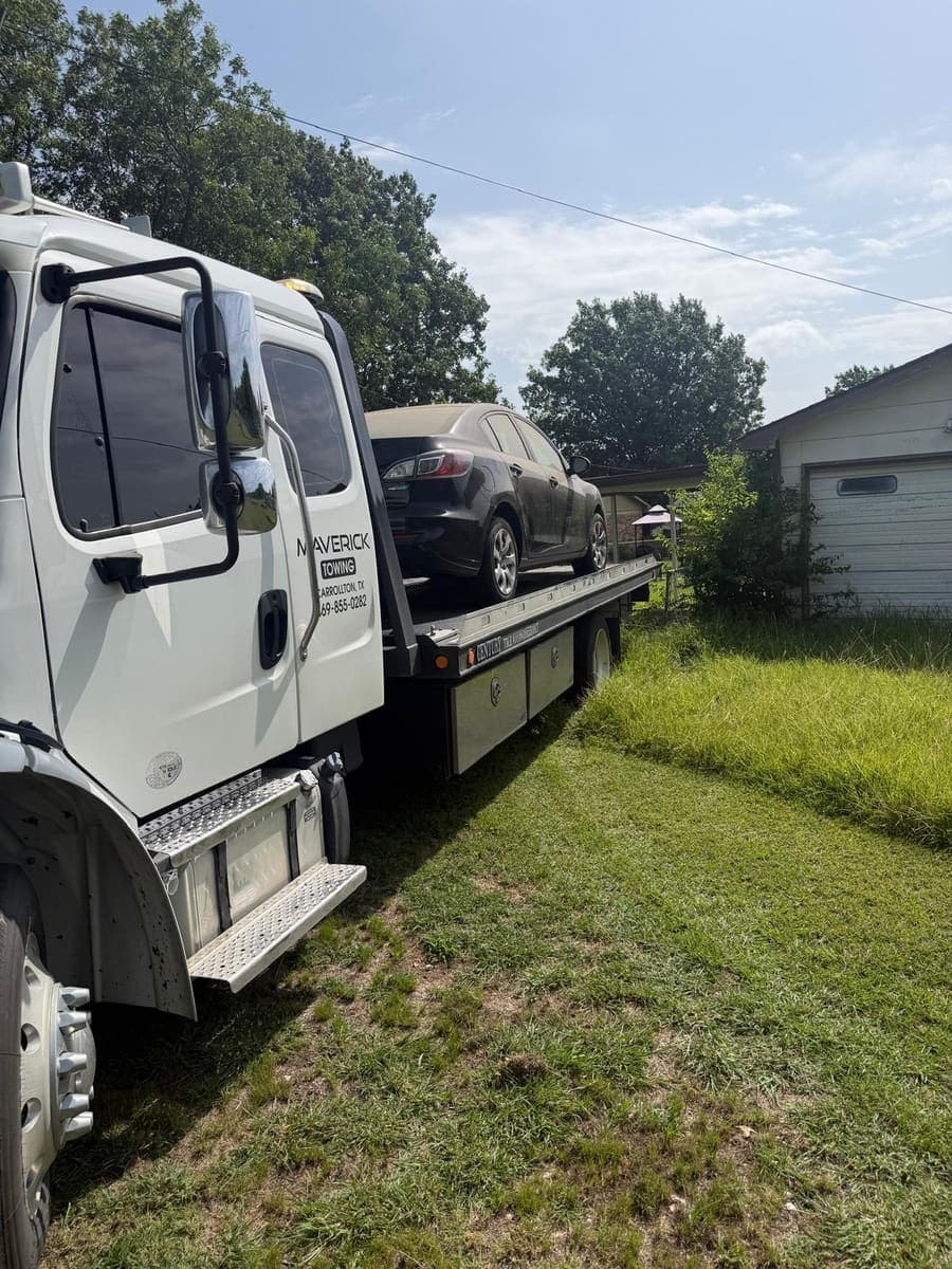 Maverick Towing flatbed hauling a black sedan out of an overgrown back yard
